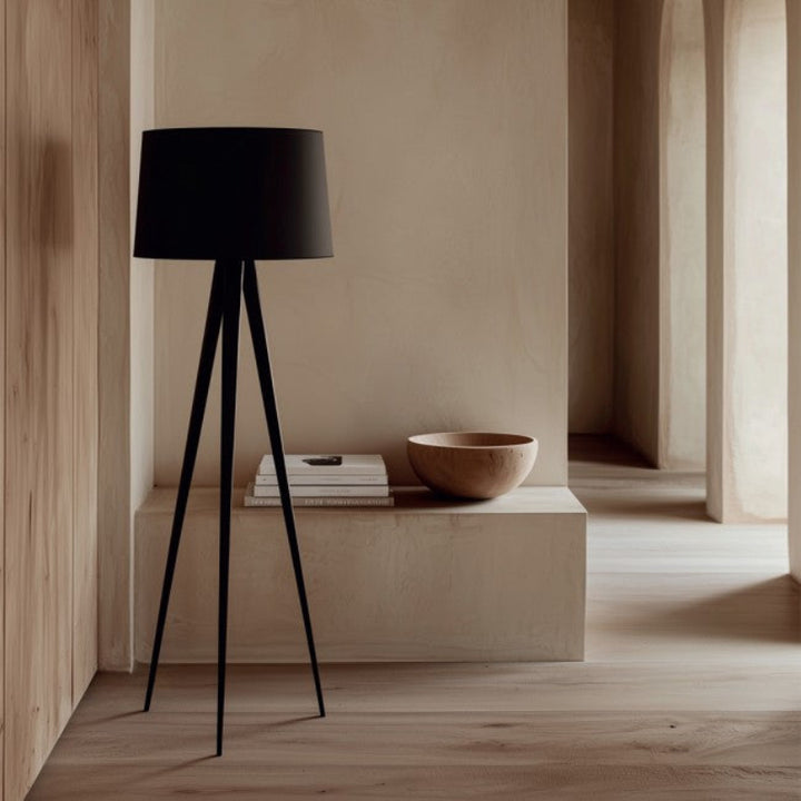 Black tripod lamp and wooden bowl on a low shelf in a minimalistic room.