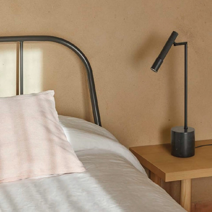 Black wall lamp on a wooden nightstand next to a bed with pink pillow and gray bedding.