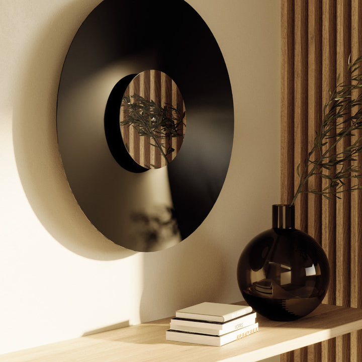 Decorative black circular mirror on a wall with a vase and books on a shelf below.